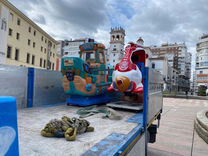Un tren de juguete y un cohete viajan en la caja de un camión en una plaza de la ciudad, con edificios al fondo.