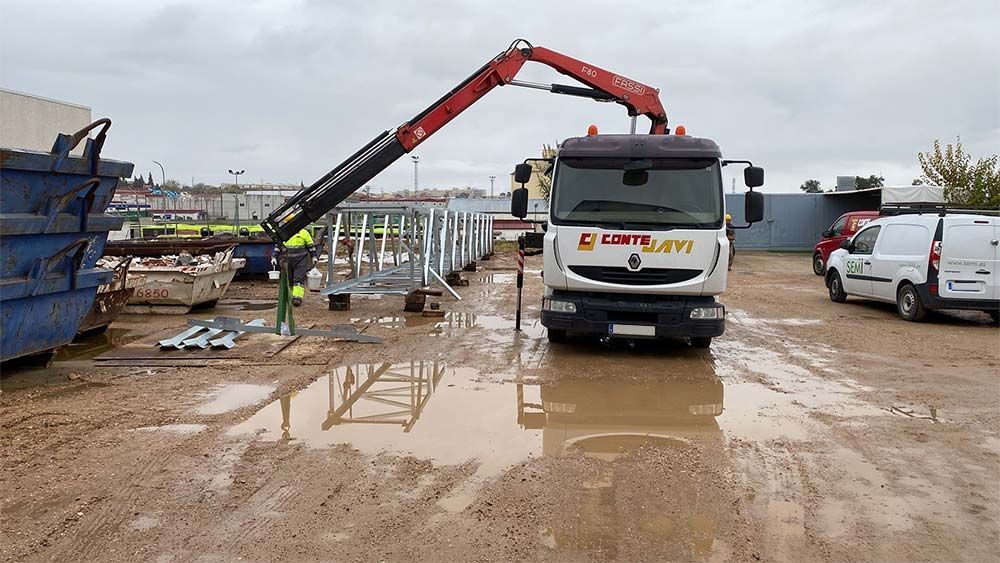 Camión con grúa levantando estructuras metálicas en una obra embarrada. También hay contenedores, trabajadores y furgonetas.