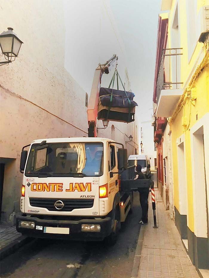 Camión blanco con grúa levantando mercancías en una calle estrecha. Una persona ayuda. Edificios flanqueados.