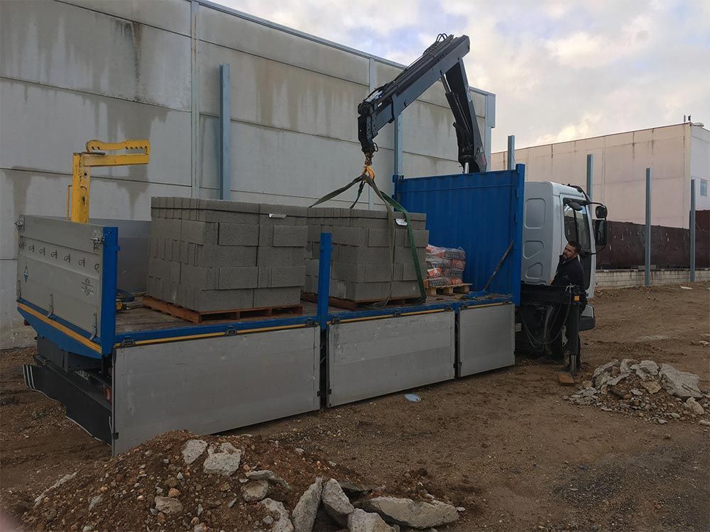 Una grúa de camión carga bloques de hormigón en un camión de plataforma cerca de un almacén.