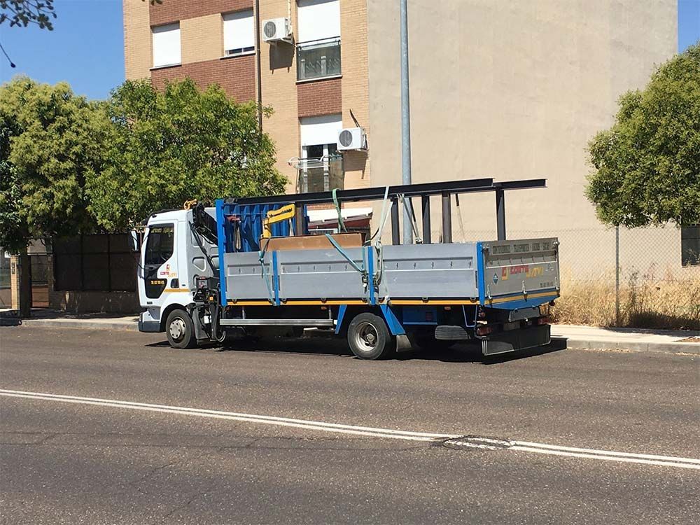 Camión con plataforma que transporta vigas metálicas, estacionado en una calle frente a un edificio.