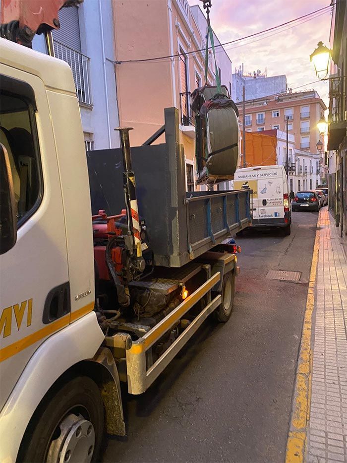 Camión cargando un contenedor en una calle estrecha al anochecer. La grúa sube un objeto a la caja del camión.