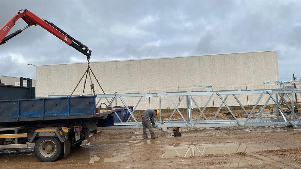 Una grúa levanta una larga estructura metálica de la caja de un camión. Un trabajador está cerca. Obra en construcción.