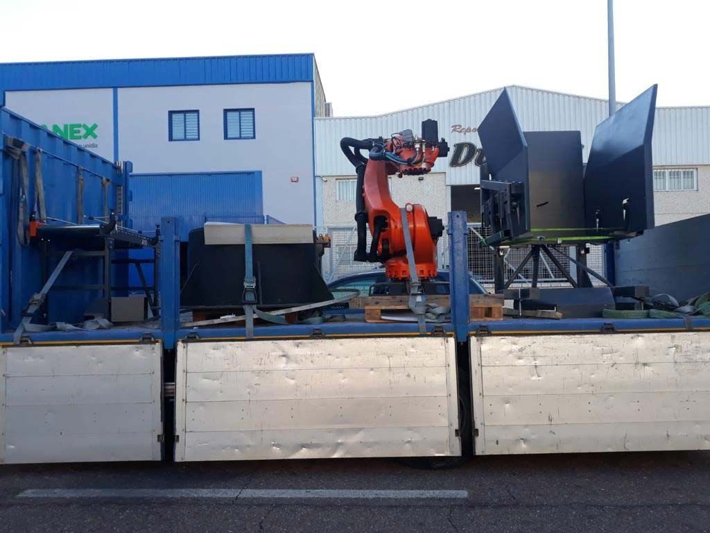 Robot industrial naranja en un camión de plataforma, equipo en el fondo, edificio azul.