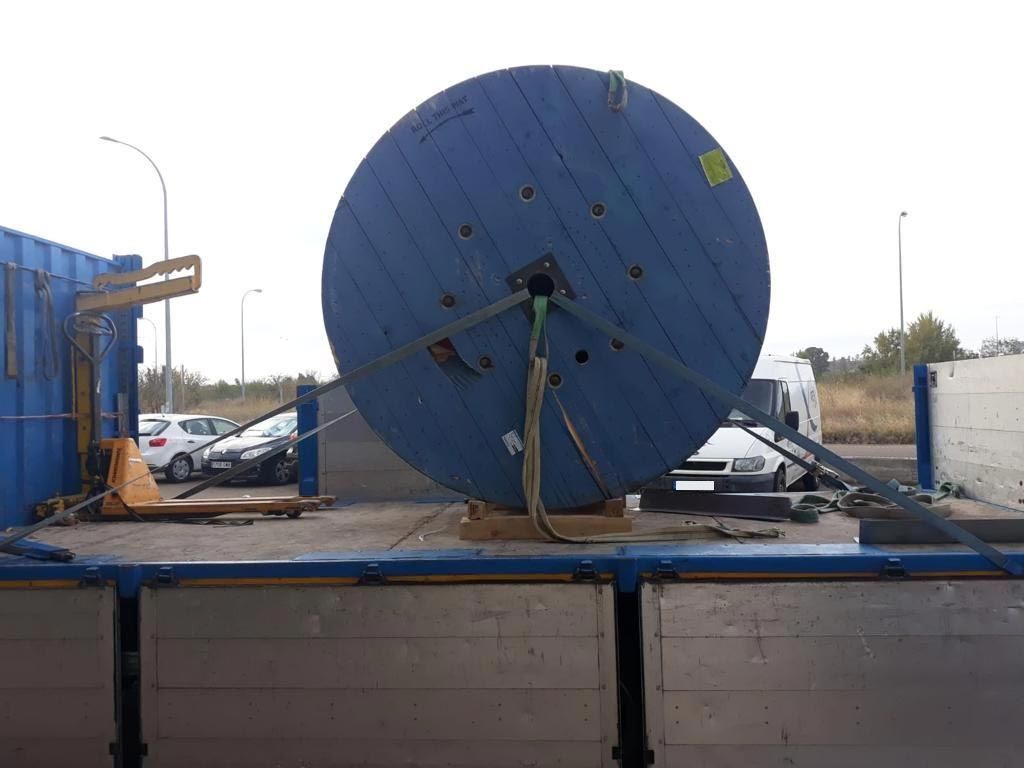 Carrete de cable azul de gran tamaño asegurado en la plataforma de un camión, sostenido por correas y una carretilla elevadora.