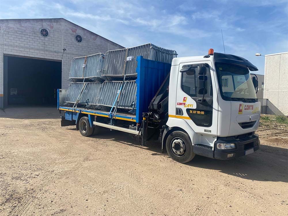 Camión blanco cargado con cercas de metal apiladas estacionado cerca de un edificio con una puerta abierta.