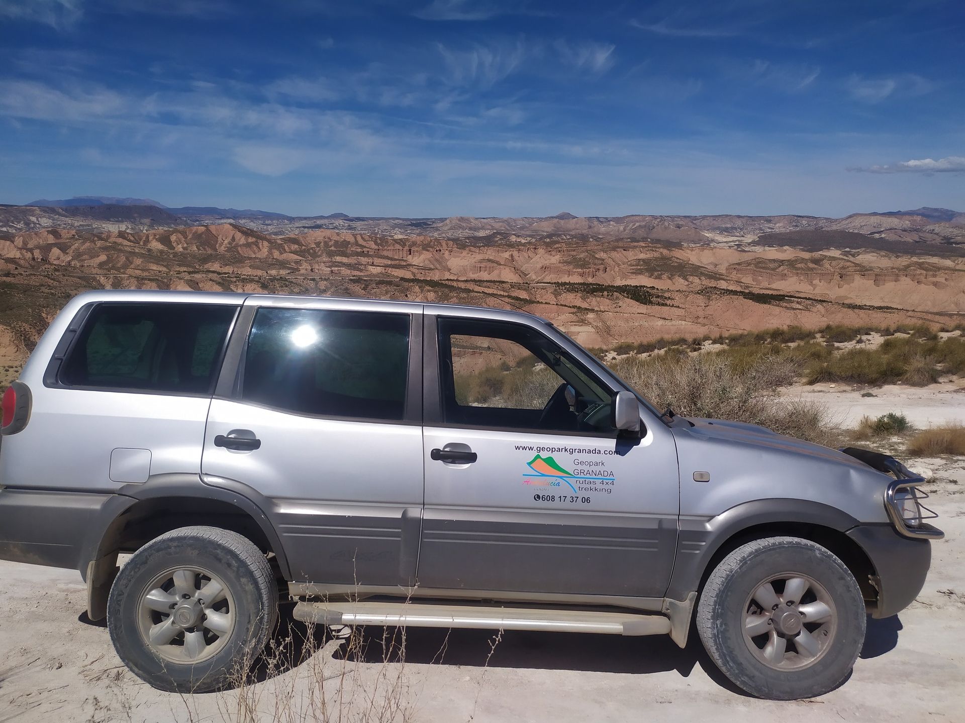 Una camioneta plateada está estacionada en un camino de tierra con montañas al fondo.