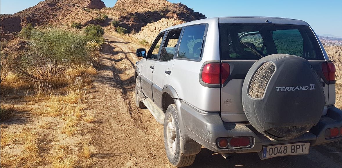 Una camioneta plateada está estacionada en un camino de tierra al lado de una montaña.