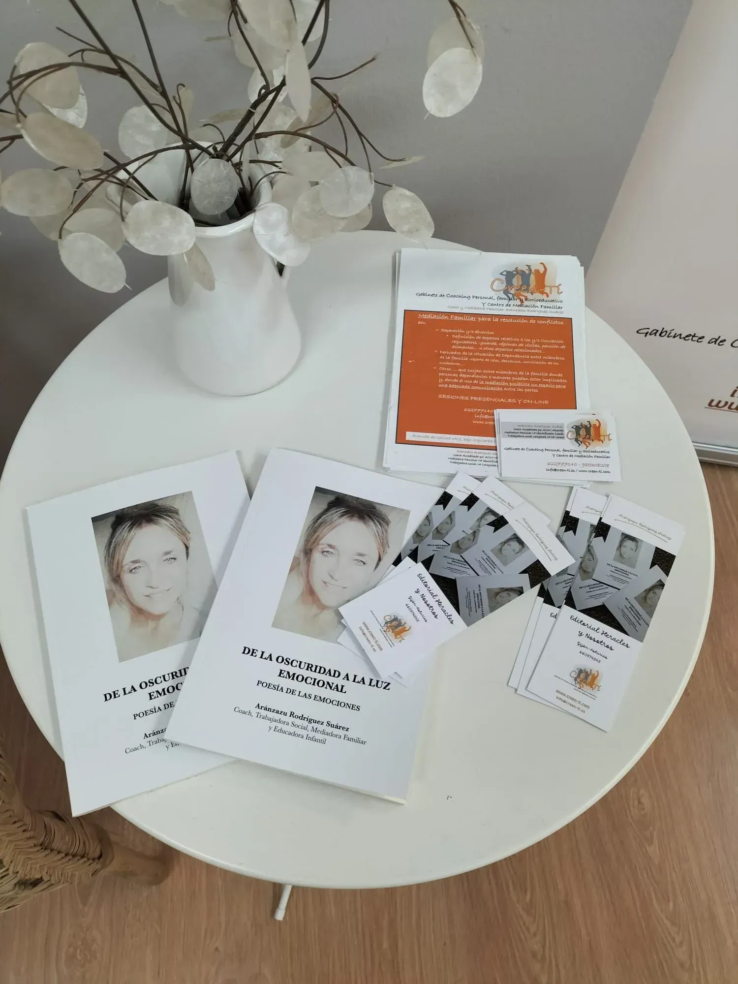 Mesa blanca con folletos, tarjetas y un jarrón con ramas decorativas blancas.