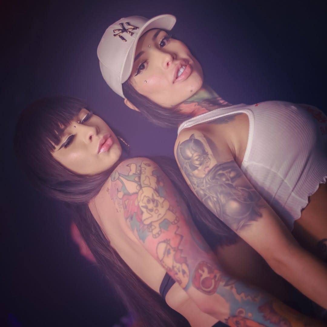 Dos mujeres tatuadas posando espalda con espalda bajo una suave luz morada, una con gorra y camiseta blanca