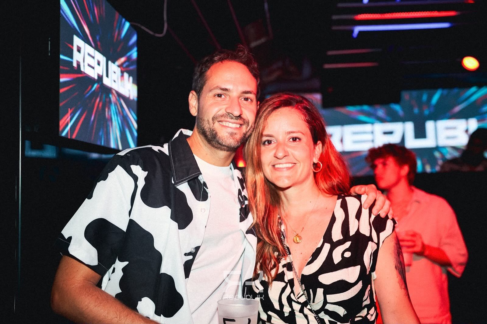 Dos personas sonríen en una discoteca iluminada con luces de neón, de pie muy juntas y mirando a la cámara.