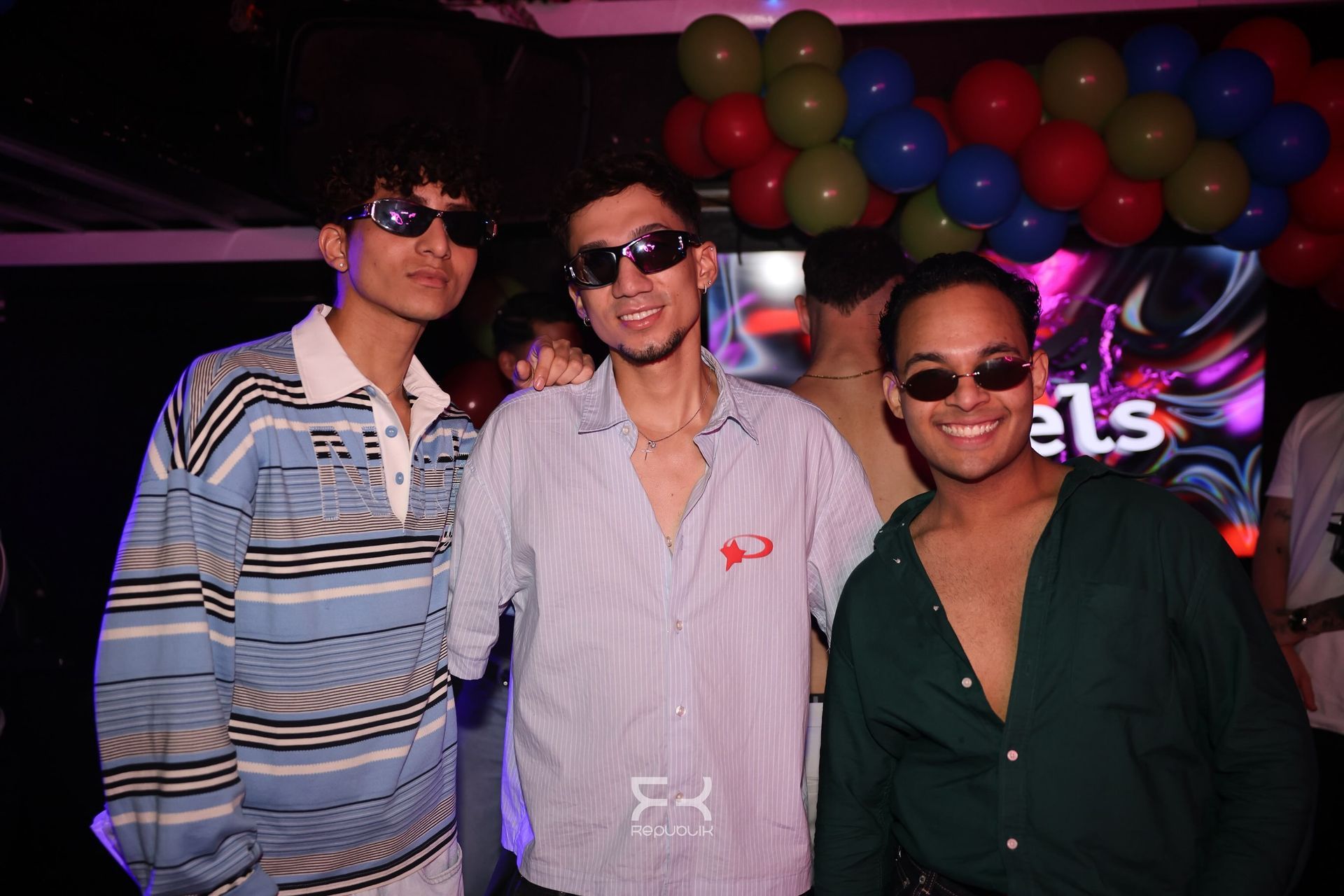 Tres personas posando en una fiesta, con globos al fondo y gafas de sol y ropa informal.