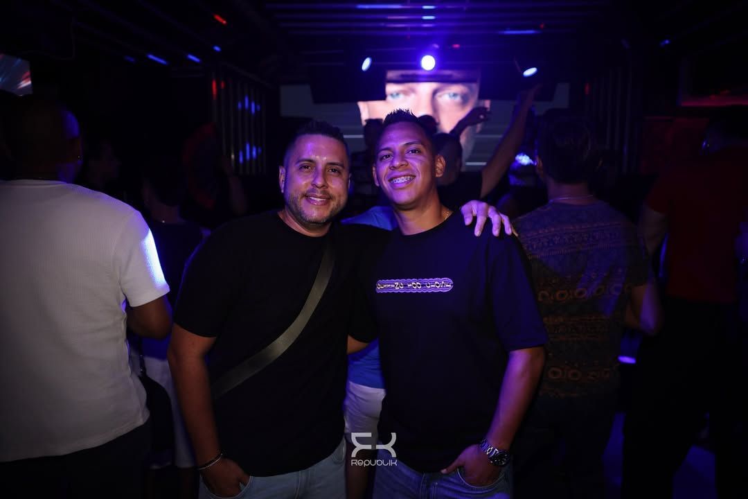 Dos personas sonriendo juntas en un club con poca luz y luces moradas.