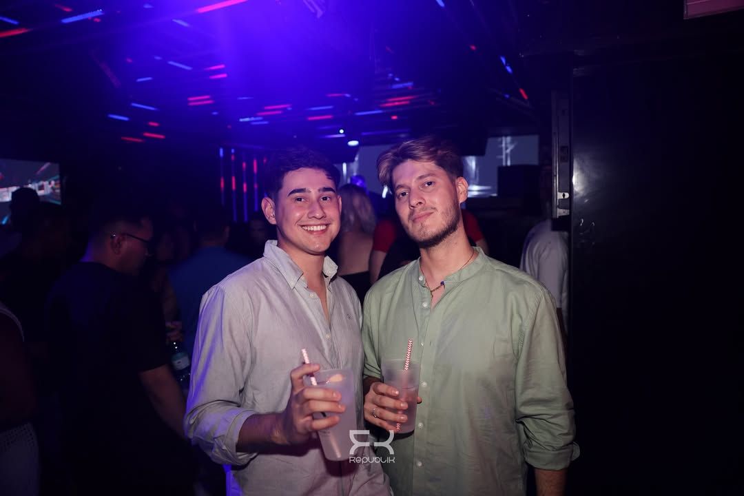 Dos personas sosteniendo bebidas y sonriendo en una discoteca con poca luz y luces azul violáceo