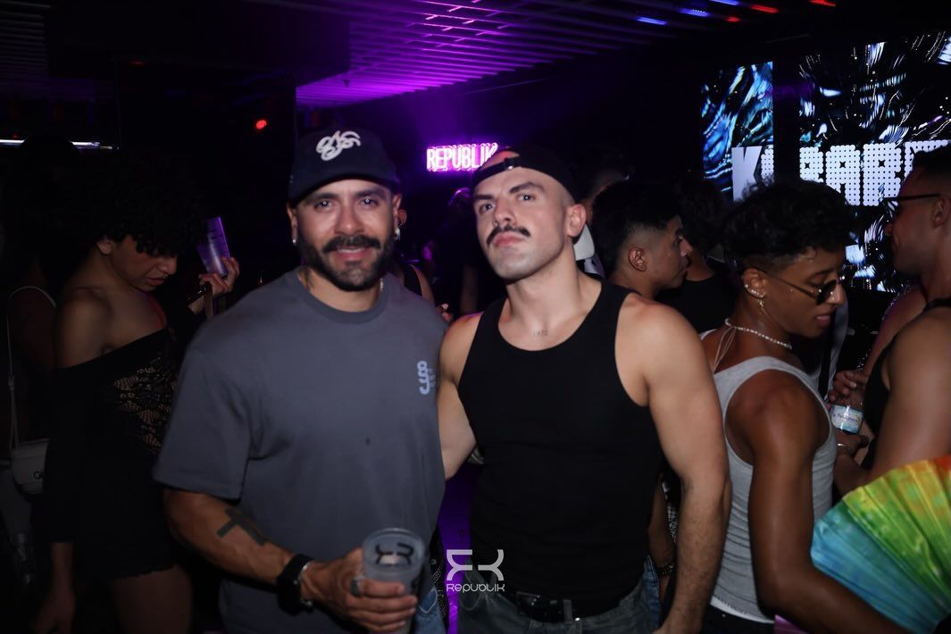 Dos hombres posando en un club con poca luz; uno sostiene una bebida bajo luces moradas.