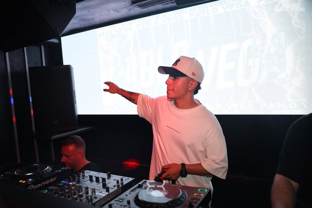 Un DJ con camisa blanca y gorra señala al público desde la cabina de una discoteca.