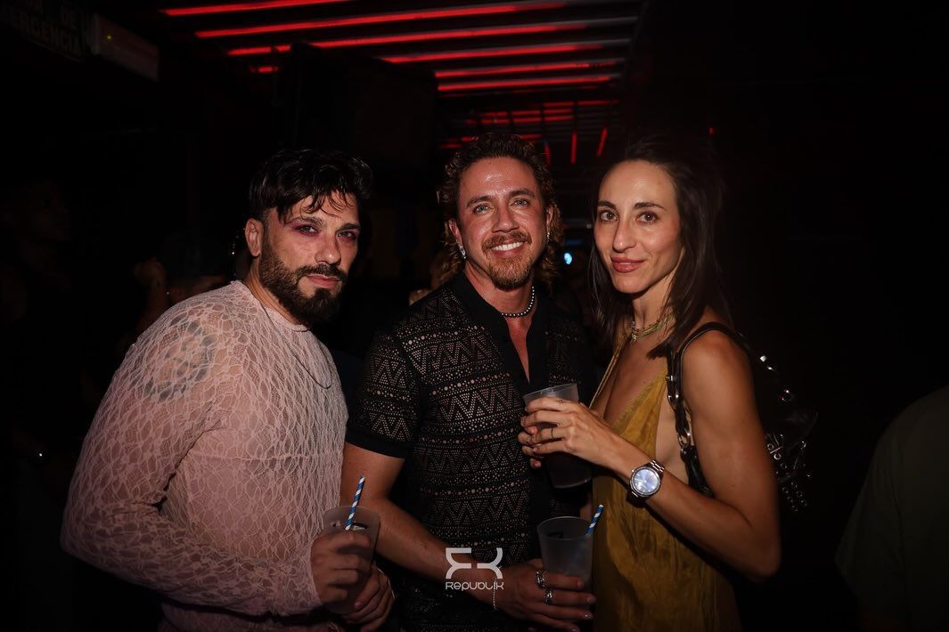 Tres personas sonríen en una fiesta con poca luz, sosteniendo bebidas bajo luces rojas.