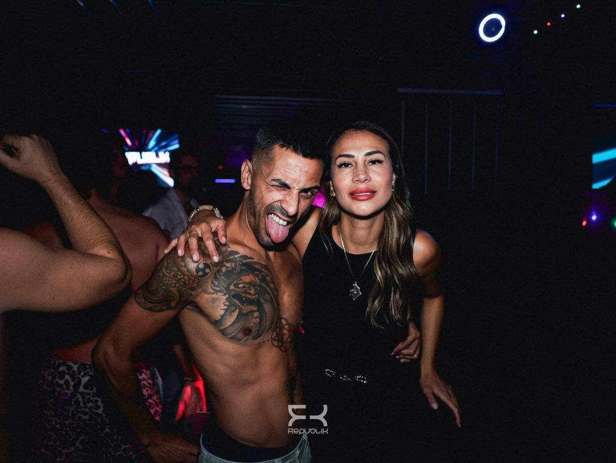 Dos personas posando en una discoteca, una sin camiseta y con tatuajes, bajo luces de colores