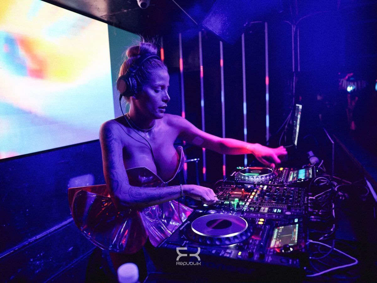 Una DJ actuando en la cabina de una discoteca, con luces de neón de colores y una gran pantalla detrás de ella