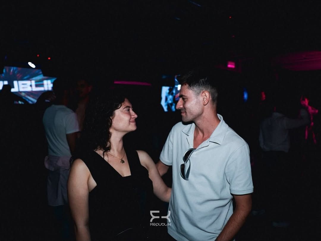 Dos personas sonríen y conversan en una discoteca poco iluminada, con luces de neón al fondo
