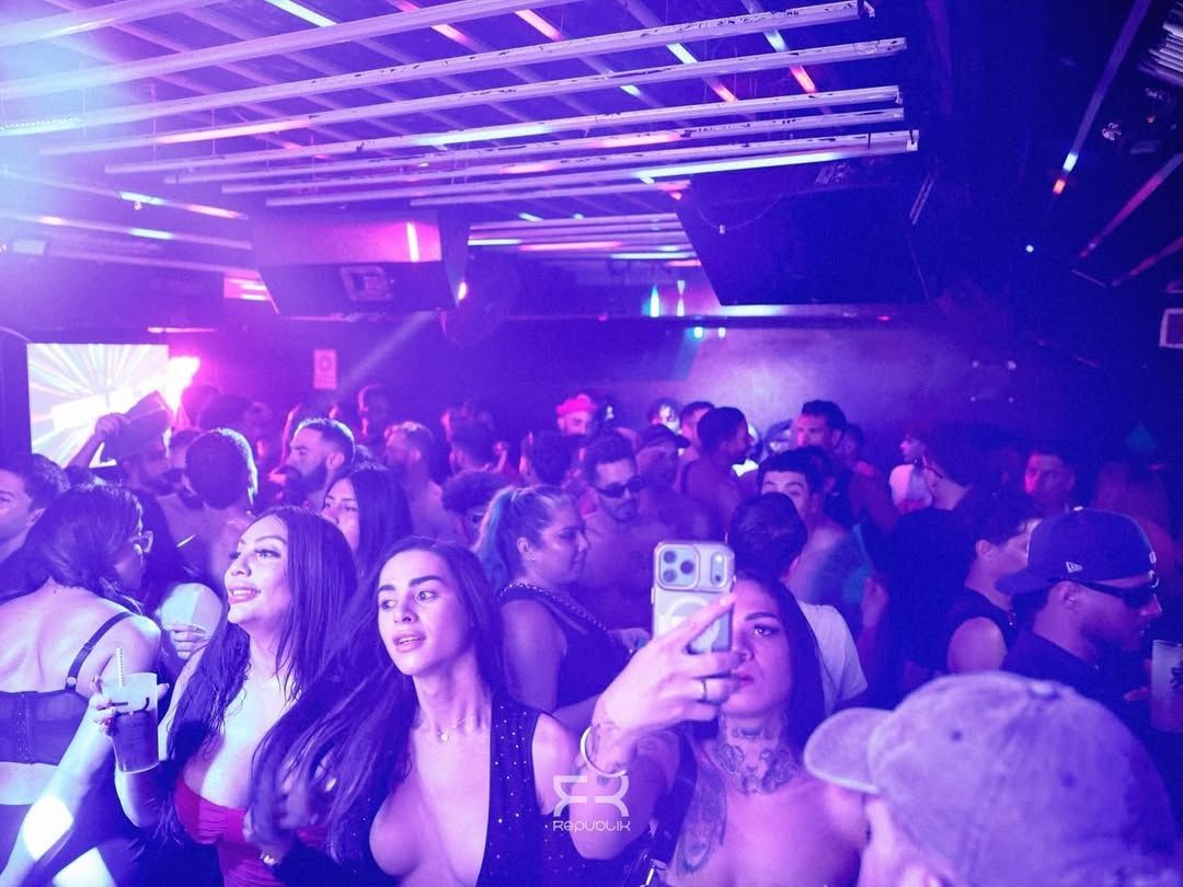 Discoteca abarrotada, con gente bailando bajo luces moradas y una mujer levantando el móvil para hacerse un selfi