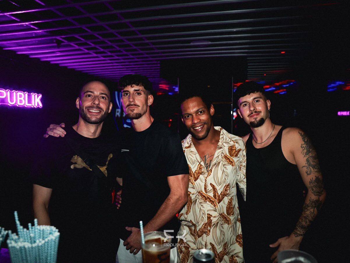 Cuatro amigos posando en una discoteca, iluminados por luces moradas y rosas, con bebidas sobre la mesa