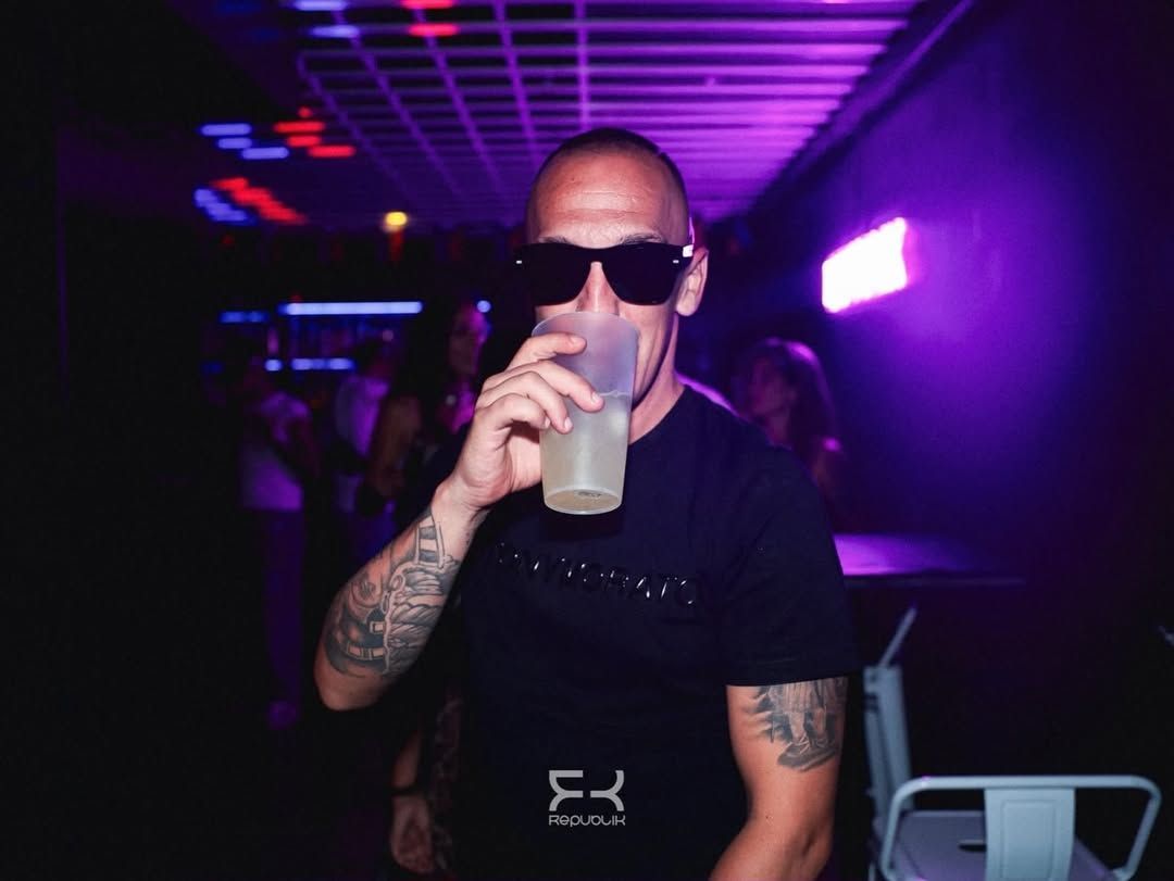 Hombre con gafas de sol sosteniendo una bebida en una discoteca iluminada con luces de neón