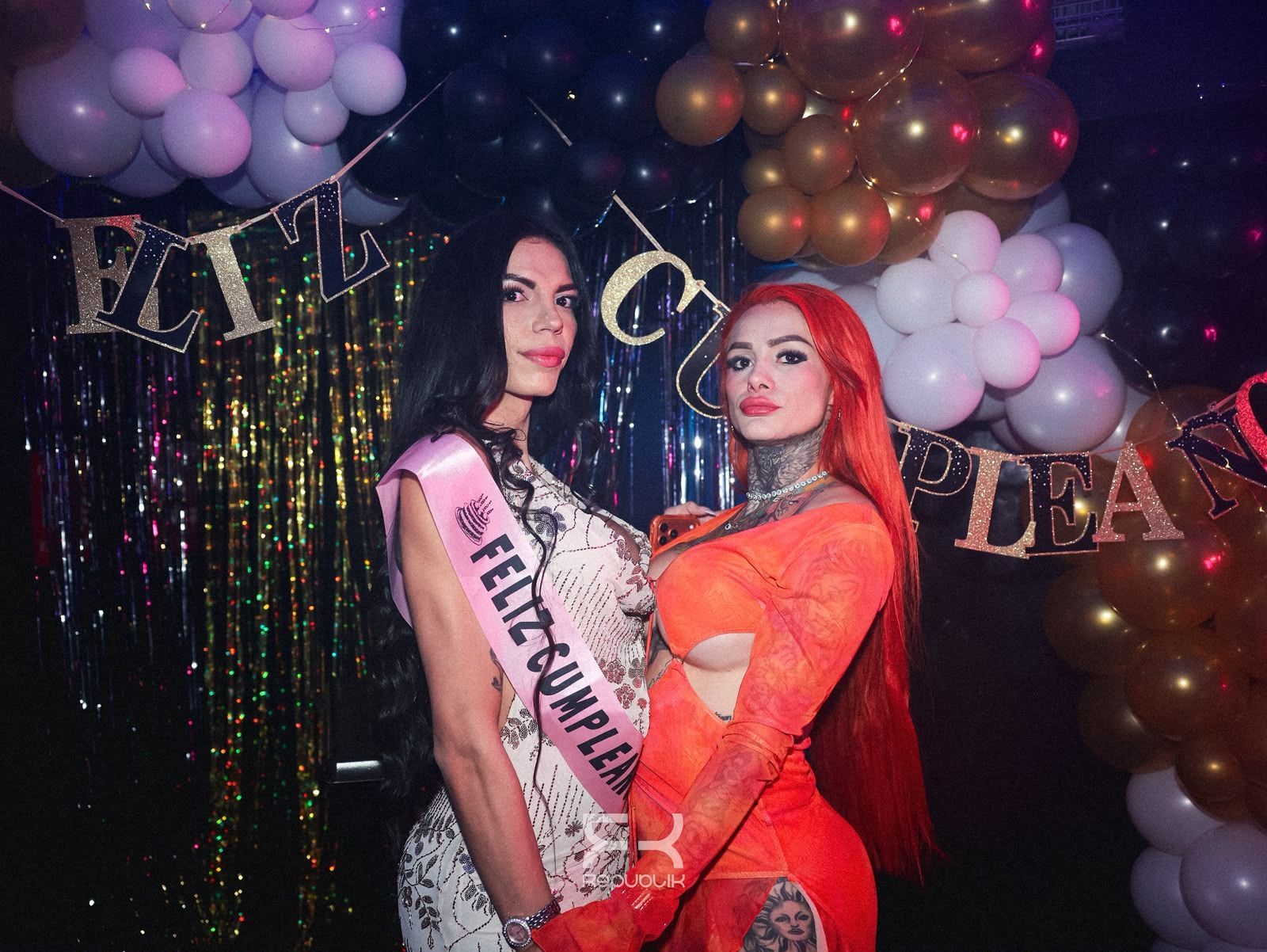 Dos mujeres con bandas de concurso de belleza posan en una fiesta, con globos y un fondo metálico
