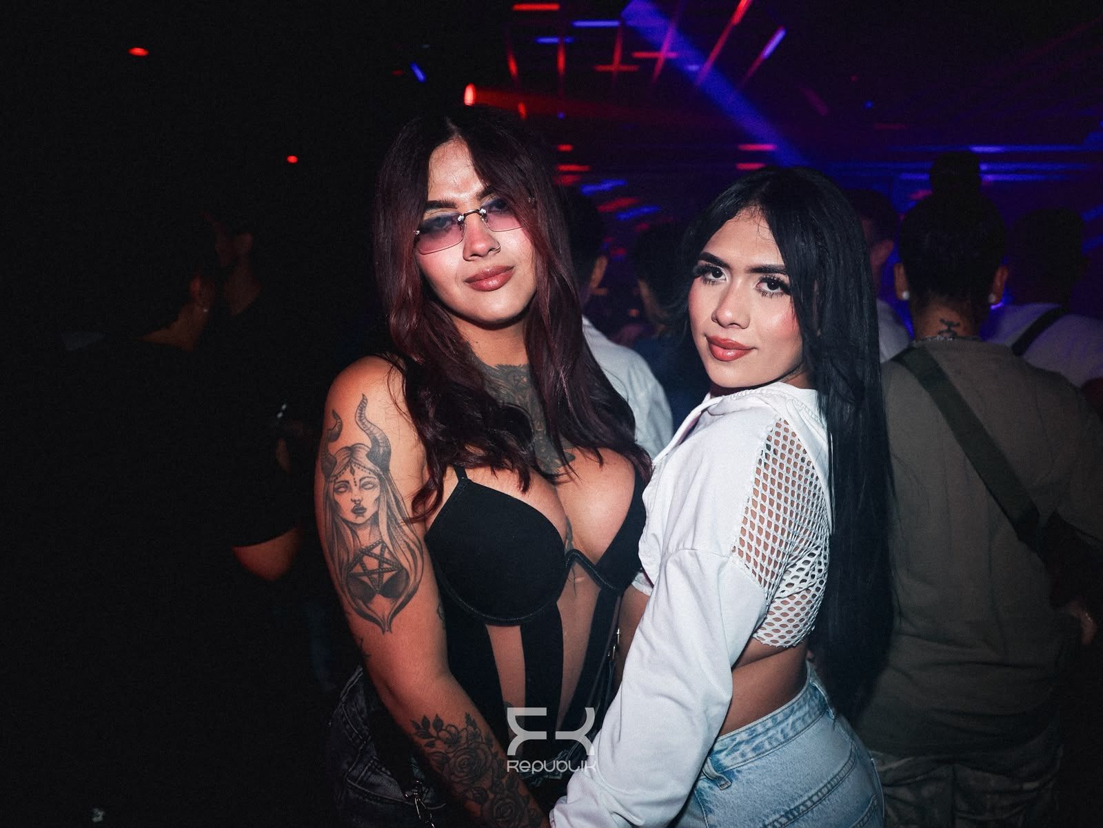 Dos mujeres posando juntas en una discoteca oscura, vestidas de blanco y negro, con luces de colores detrás de ellas