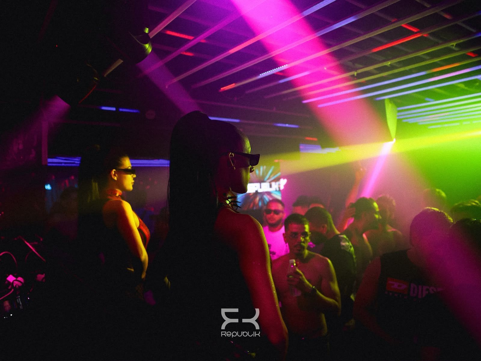 Discoteca abarrotada, con gente bailando bajo luces láser rosas, verdes y amarillas