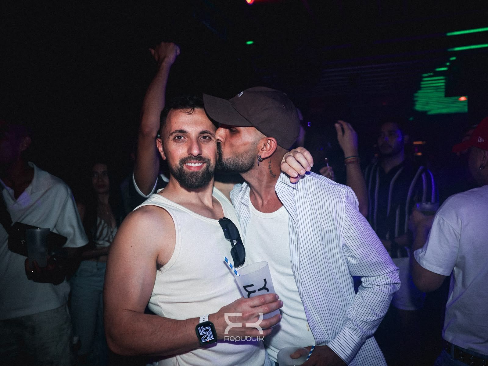 Dos hombres posando en un club con poca luz; uno sostiene una bebida y, al fondo, hay luces de colores