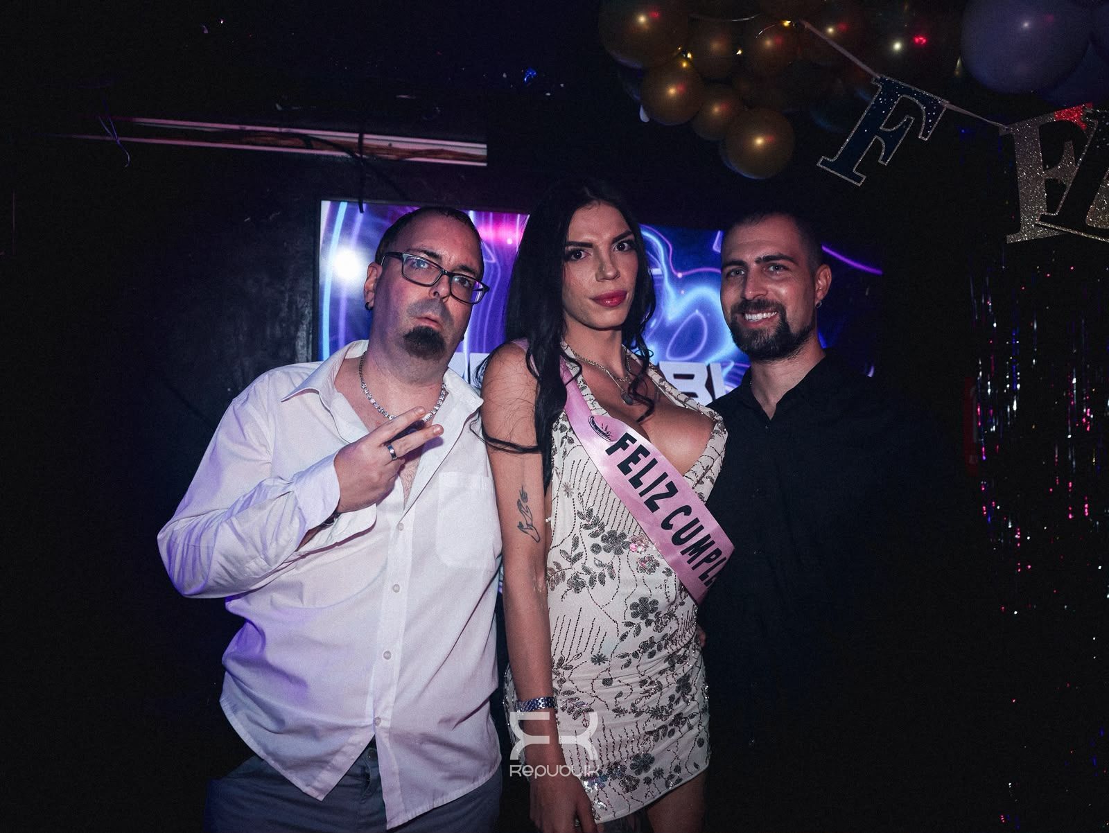 Tres personas posando en una fiesta, con globos y poca luz al fondo.