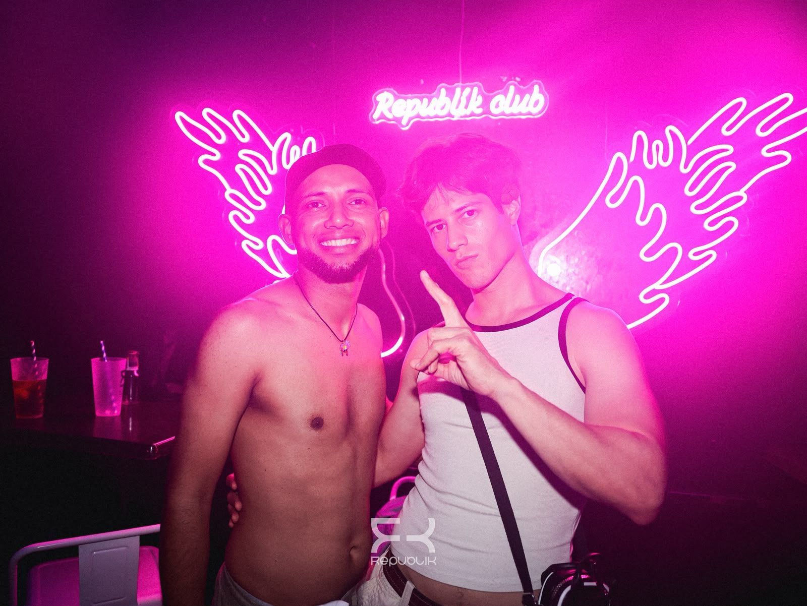 Dos hombres posando en un club rosa neón con alas de ángel en la pared, uno sin camiseta y el otro con una camiseta de tirantes blanca