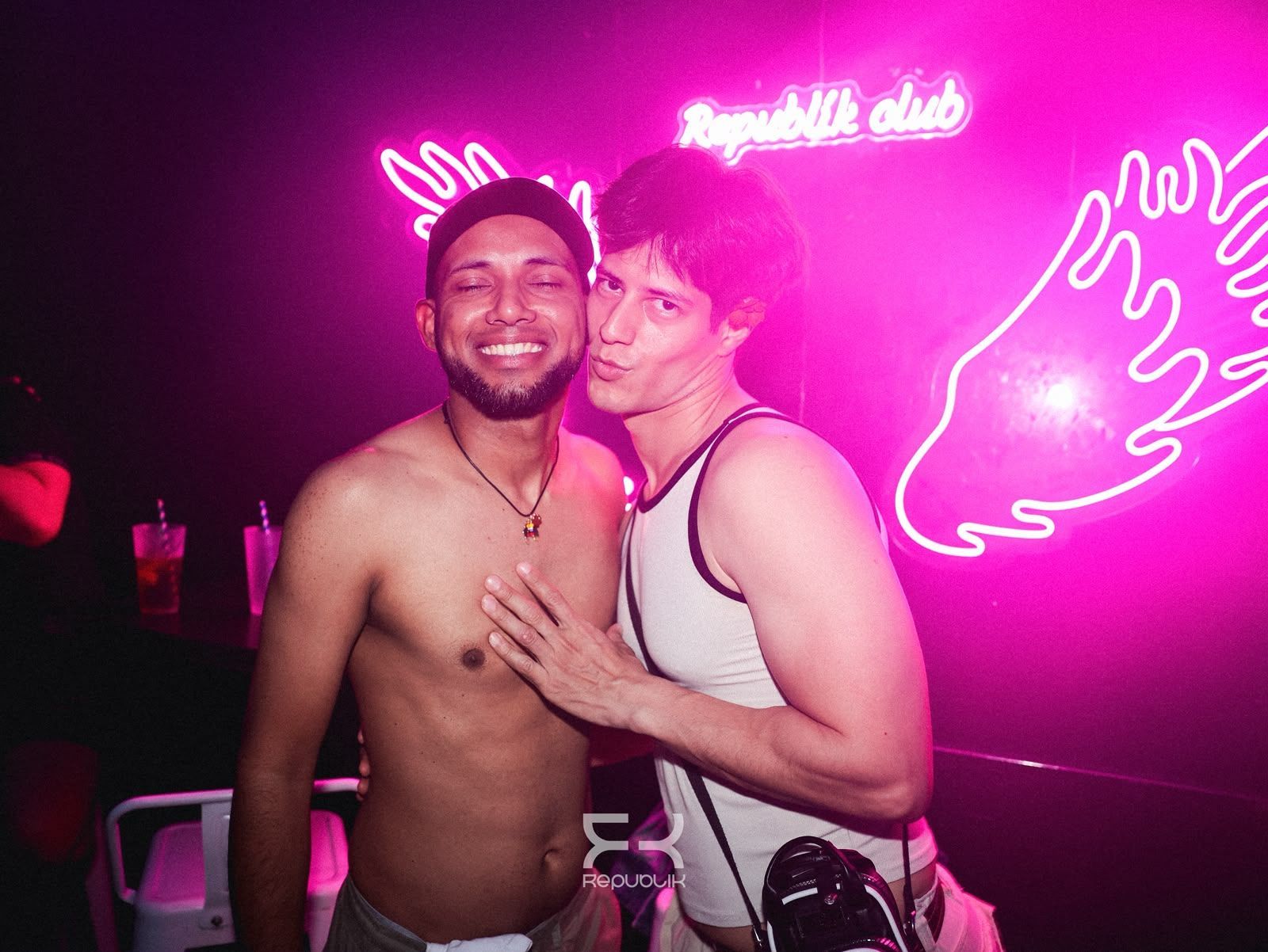 Dos personas posando juntas en un club iluminado con luces de neón rosas y decorado con arte mural en forma de alas