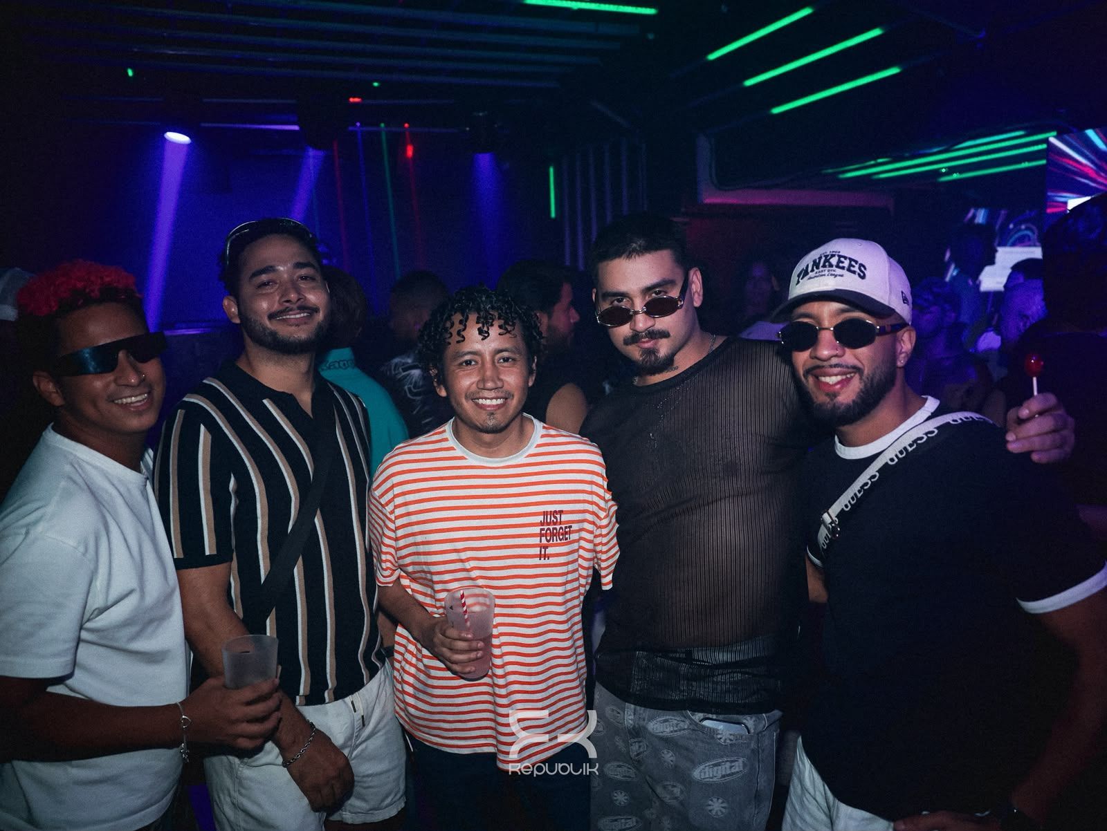 Cinco amigos posan juntos en una discoteca poco iluminada, con luces de neón, y sonríen a la cámara.