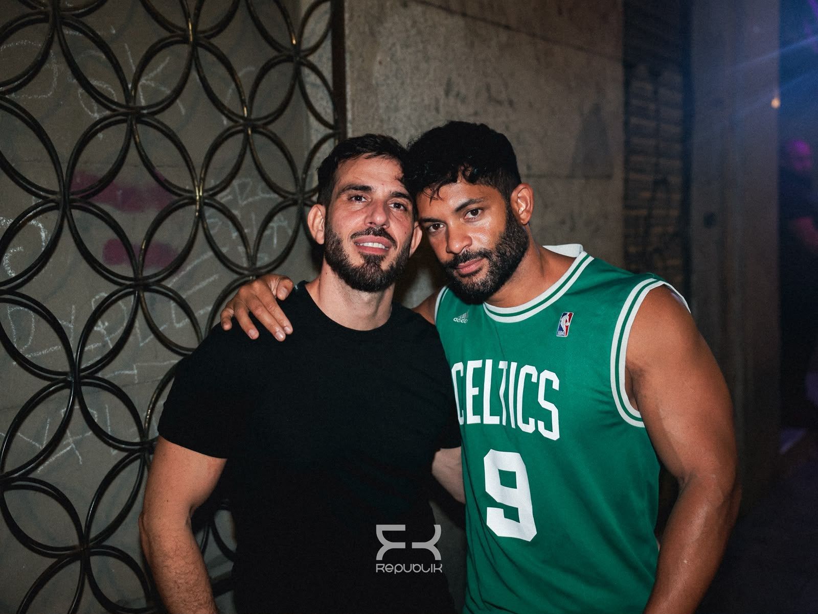 Dos hombres posan juntos en un espacio interior: uno lleva una camiseta negra y el otro una camiseta verde de los Celtics.