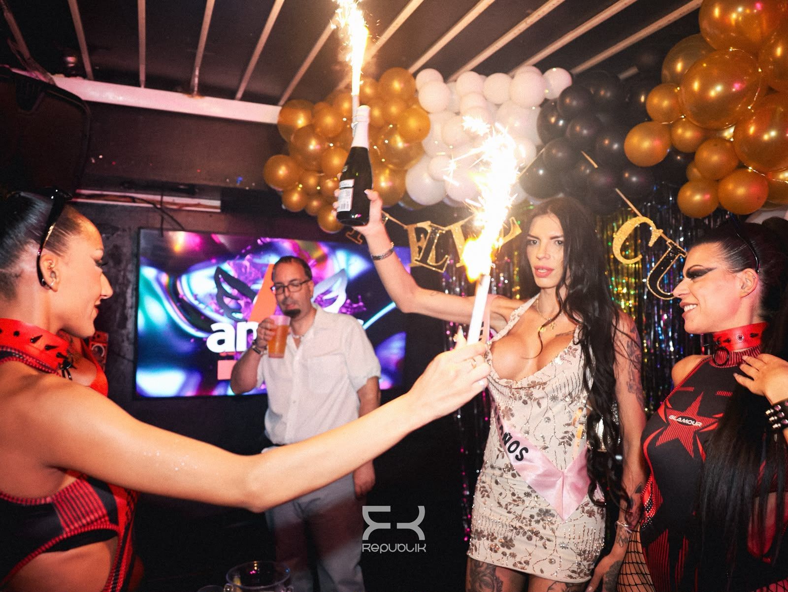 Personas bailando en una fiesta con globos y un fondo iluminado; una de ellas sostiene una botella de champán en la pista de baile