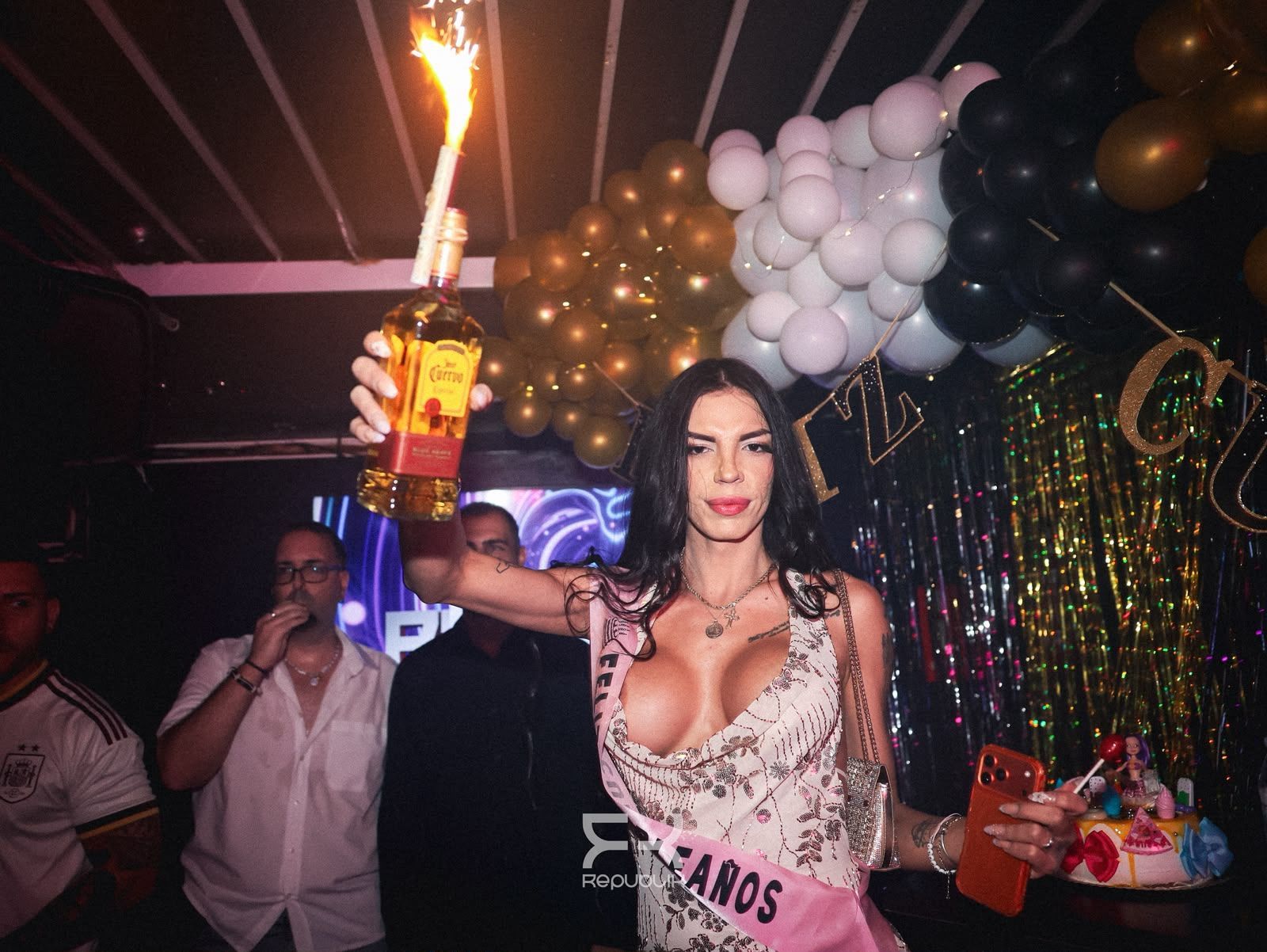 Una persona en una fiesta sostiene una botella en llamas junto a unos globos y una colorida mesa de postres