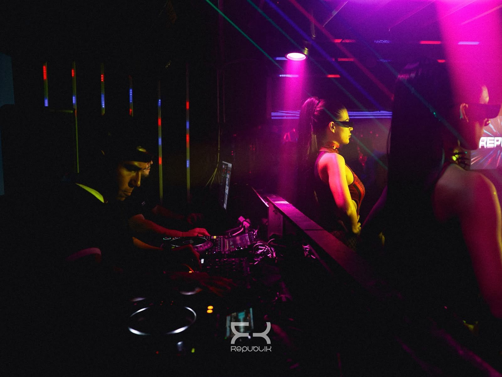 Cabina de DJ en un club oscuro, iluminada por luces de neón rosas y moradas, con gente bailando cerca