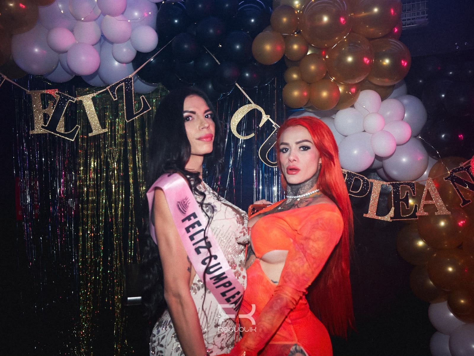 Dos mujeres posan en una fiesta delante de una decoración de globos dorados y blancos; una lleva una banda.