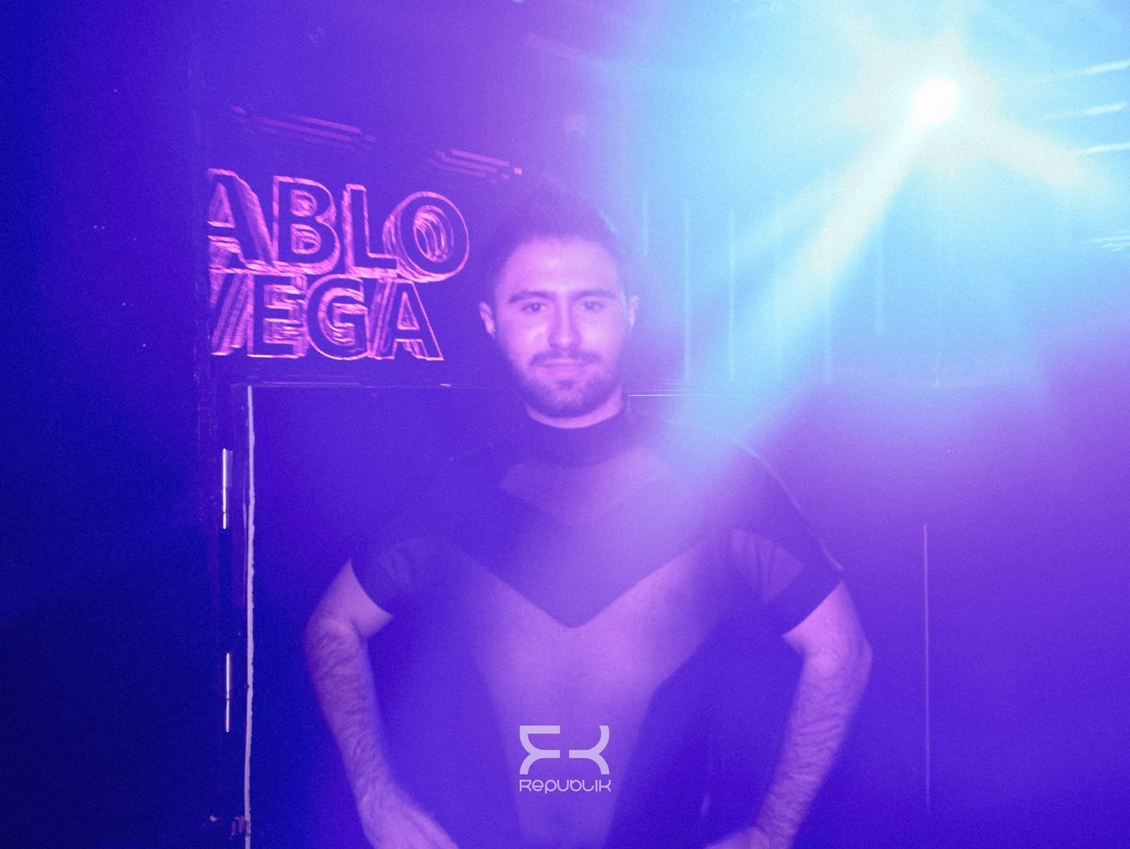 Hombre de pie en un escenario iluminado con luces de neón moradas y un intenso foco azul sobre él