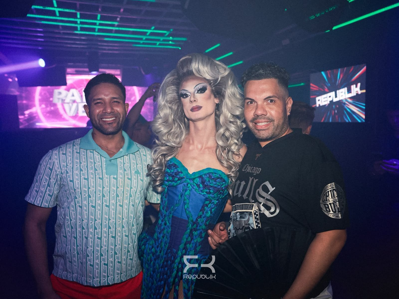Tres personas posando en una discoteca iluminada con luces de neón, con un escenario de fondo y luces de colores detrás