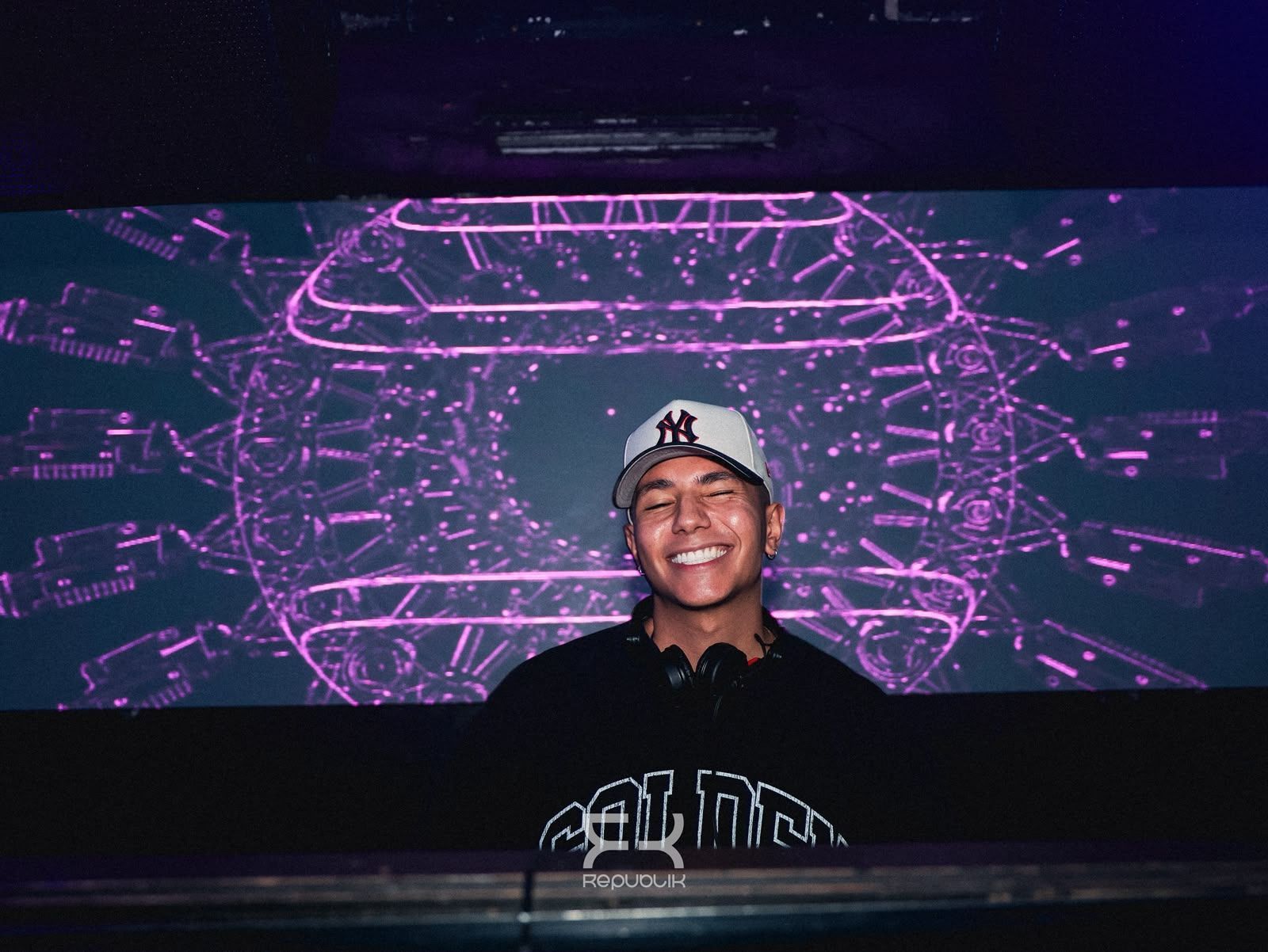Una persona sonriente con gorra está de pie ante un fondo geométrico de neón morado.