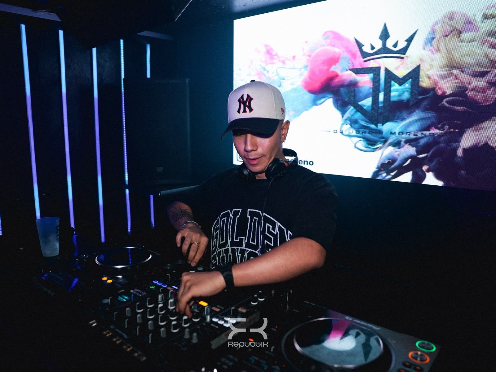 Un DJ mezclando con una tornamesa en un club iluminado con luces de neón, con un fondo de logotipos coloridos detrás de él.