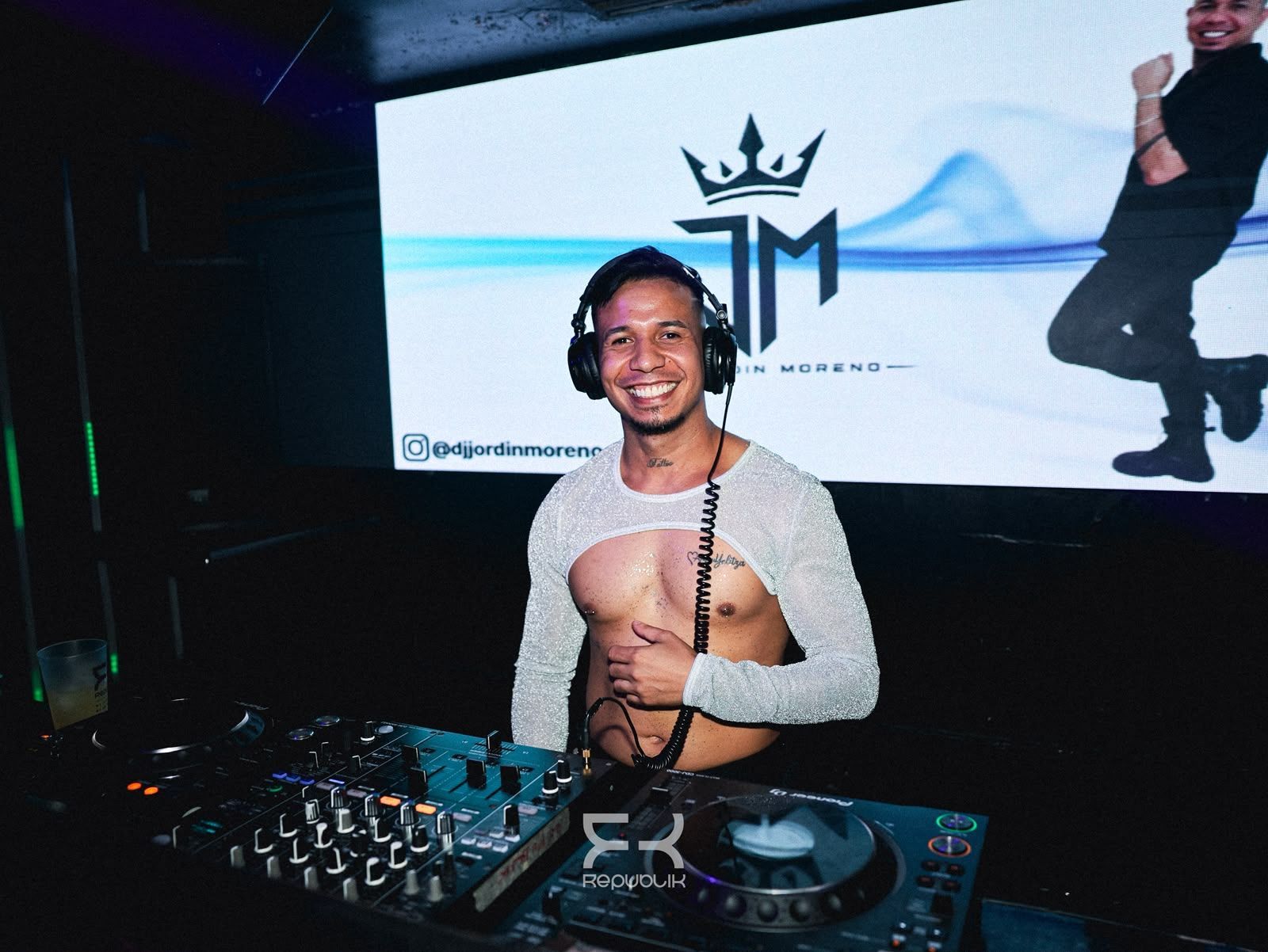 DJ sonriente detrás de una mesa de mezclas en la cabina de una discoteca, con una pantalla iluminada al fondo