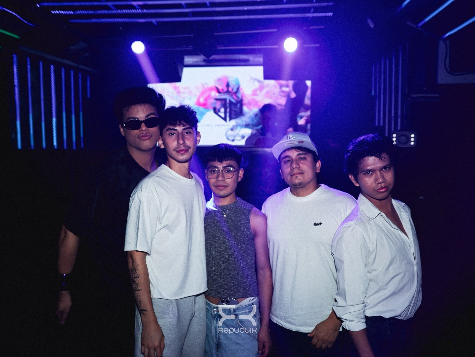 Cinco personas posando en una discoteca iluminada de color morado, con un escenario y una pantalla detrás.