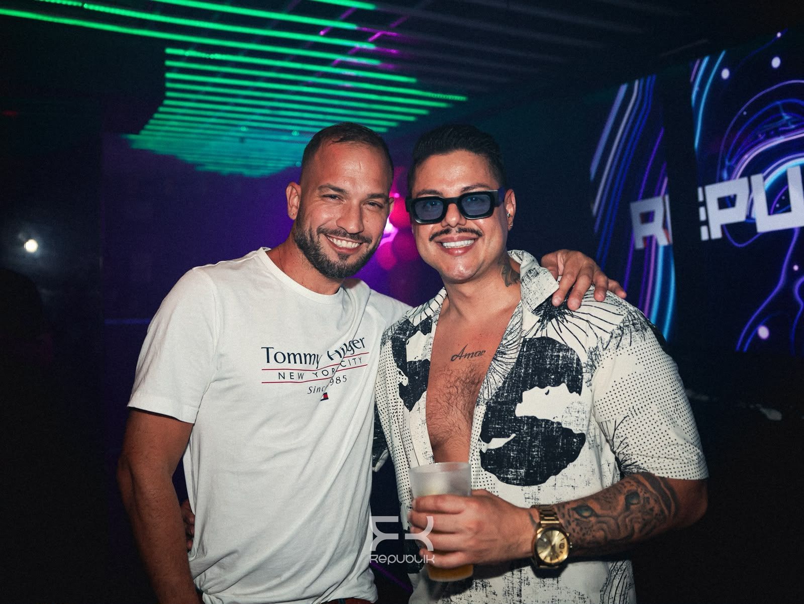 Dos hombres posan sonrientes en una discoteca iluminada con luces de neón, uno de ellos sosteniendo una bebida.