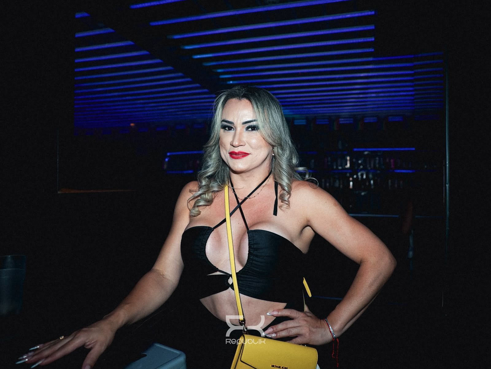 Una mujer posando con un vestido negro bajo luces de neón azules en un local con poca iluminación