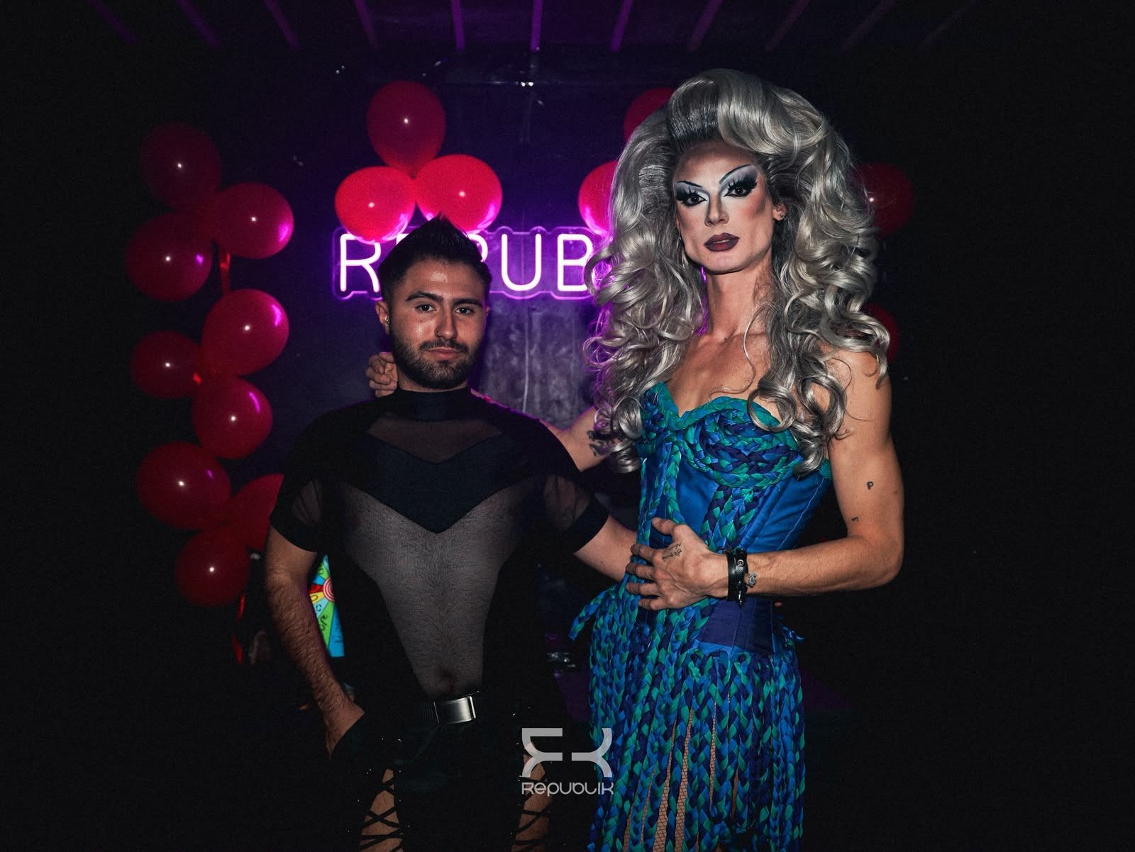 Dos personas posando frente a un fondo morado con globos rojos en un evento en una discoteca