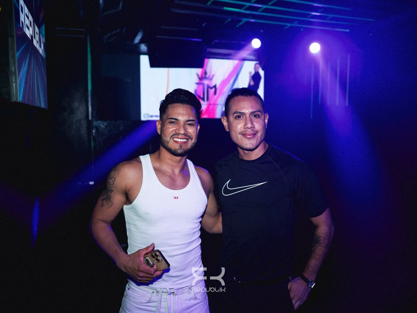 Dos hombres posando juntos en una discoteca con poca iluminación y luces moradas en el escenario